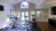 This fitness center is designed to inspire an active lifestyle, combining thoughtful architecture with a comprehensive selection of exercise equipment. The space is defined by its dramatic vaulted ceilings that rise to a striking arched window at the peak, flooding the room with natural light and creating an airy, expansive atmosphere. A ceiling fan mounted beneath the arch provides comfortable airflow throughout workouts.

The workout area is outfitted with a variety of Life Fitness cardio machines, including treadmills and an elliptical or stair-stepping unit, all positioned on protective rubber flooring that covers a generous portion of the space. Additional strength and multi-use training equipment is arranged throughout the room, offering a well-rounded fitness experience.

Large French doors open to an outdoor area, further enhancing the sense of openness and allowing for natural ventilation. Flanking the doors, wide windows with horizontal blinds admit additional daylight while maintaining privacy. The walls are finished in a neutral tone that complements the dark hardwood-style flooring bordering the rubber mat area. A motivational fitness poster adorns one wall, adding an energetic touch. Overall, the space strikes a balance between functionality and an inviting, light-filled environment.