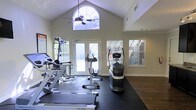 This fitness center is designed to inspire an active lifestyle, combining thoughtful architecture with a comprehensive selection of exercise equipment. The space is defined by its dramatic vaulted ceilings that rise to a striking arched window at the peak, flooding the room with natural light and creating an airy, expansive atmosphere. A ceiling fan mounted beneath the arch provides comfortable airflow throughout workouts.

The workout area is outfitted with a variety of Life Fitness cardio machines, including treadmills and an elliptical or stair-stepping unit, all positioned on protective rubber flooring that covers a generous portion of the space. Additional strength and multi-use training equipment is arranged throughout the room, offering a well-rounded fitness experience.

Large French doors open to an outdoor area, further enhancing the sense of openness and allowing for natural ventilation. Flanking the doors, wide windows with horizontal blinds admit additional daylight while maintaining privacy. The walls are finished in a neutral tone that complements the dark hardwood-style flooring bordering the rubber mat area. A motivational fitness poster adorns one wall, adding an energetic touch. Overall, the space strikes a balance between functionality and an inviting, light-filled environment.