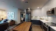 Located at 18418 Dearborn St in the Northridge neighborhood of Los Angeles, this single-floor unit offers a thoughtfully designed interior with a cohesive modern aesthetic. The open-concept kitchen is a standout feature, showcasing two-tone cabinetry with dark lower cabinets and crisp white upper cabinets, a decorative tile backsplash, stainless steel appliances including a full-size refrigerator, dishwasher, range, and over-the-range microwave, and clean white countertops. A rustic wood dining table sits adjacent to the kitchen, providing a warm contrast to the contemporary finishes. Hardwood-style flooring runs throughout the kitchen and dining area. The living room features horizontally striped accent walls in cool grey tones and opens directly to a private balcony through large sliding glass doors, allowing natural light to fill the space. The bedroom is designed with a bold geometric accent wall in navy, teal, and grey, adding a strong visual focal point, and is finished with plush carpeting. A frosted window provides natural light while maintaining privacy. The bathroom is well-appointed with a dark vanity, white countertop, illuminated LED mirror, floating wood shelves with black industrial brackets, and wood-tone flooring, delivering a polished and functional finish.