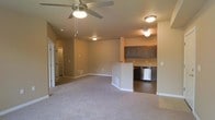 This single-floor space offers a thoughtfully designed open-concept layout that seamlessly integrates the living area and kitchen. The living space is finished with plush, neutral-toned carpeting that adds warmth and comfort underfoot, while a ceiling fan with integrated lighting provides both airflow and ambient illumination.

The kitchen is defined by dark-toned cabinetry that contrasts elegantly with the lighter walls, and features stainless steel appliances, including a dishwasher. A peninsula-style breakfast bar separates the kitchen from the living area, offering a functional space for casual dining or meal preparation. The kitchen flooring transitions to a hard surface, providing a practical and easy-to-clean workspace.

Flush-mount ceiling fixtures are distributed throughout the space, ensuring consistent, even lighting across all areas. A hallway visible from the main living space leads to additional rooms, suggesting a well-organized floor plan. The warm, neutral color palette applied throughout the walls ties the spaces together cohesively, creating a welcoming and functional environment suitable for everyday living.