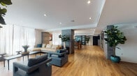 This thoughtfully designed single-floor amenity space offers an expansive, open-plan layout united by warm wide-plank hardwood flooring and recessed lighting throughout. The lounge area serves as the social centerpiece, furnished with a generous sectional sofa and coordinating blue upholstered armchairs arranged around a sleek coffee table. Floor-to-ceiling sheer curtains line the exterior walls, allowing abundant natural light to fill the space while softening the overall ambiance.

A striking dark-finished island with a polished countertop anchors the wet bar area, accommodating multiple bar stools with brass-accented legs. Behind the island, custom slate-blue cabinetry with brass hardware frames a vibrant teal subway tile backsplash, built-in open shelving, and an integrated sink — creating a functional yet visually compelling focal point.

The game and entertainment zone features a full-size billiards table as its centerpiece, complemented by a wall-mounted flat-screen television and a small seating arrangement ideal for casual gatherings. Bold monochromatic artwork and textured accent walls add artistic depth to this area. Structural columns clad in dark stone-effect material provide architectural definition between zones. The space is accessible via glass-paned double doors, reinforcing its open, communal character.