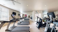 Located at 4097 N Cadillac Dr in Fayetteville, Arkansas, this fitness center offers a generously sized, single-floor workout facility designed to support a full range of exercise routines. The open layout is bright and welcoming, with large windows along the perimeter allowing ample natural light to fill the space and provide views of the surrounding exterior.

The cardio zone features a row of TRUE-brand treadmills and elliptical trainers, as well as upright and recumbent bikes, each equipped with personal display screens. Ceiling fans are positioned throughout to maintain airflow and comfort during workouts. Wall-mounted televisions add an additional entertainment option for cardio sessions.

The strength training area includes a multi-station cable machine by Cybex, a weight bench, and a full dumbbell rack reflected by a floor-to-ceiling mirror wall, which enhances the sense of openness. Stability balls are available for core and flexibility work.

The space is unified by a spirited University of Arkansas Razorbacks theme, with the iconic Razorback logo prominently displayed and motivational wall decals reading phrases such as "Sore Today, Strong Tomorrow," "Pig Sooie," and "Believe in Yourself," lending the facility an energetic and community-driven atmosphere. Recessed lighting and neutral-toned carpet flooring complete the clean, functional aesthetic of this well-appointed fitness space.