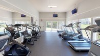 This fitness center is a thoughtfully designed workout facility that offers a comprehensive range of exercise equipment within a bright, open environment. The space features soaring ceilings that lend an airy, open feel, while large sliding glass doors and generously sized windows flood the room with natural light and provide pleasant views of the surrounding greenery outdoors.

The cardio section is well-stocked with multiple treadmills, elliptical trainers, and a stair climber, all arranged to take advantage of the natural light and outdoor views. For strength training, the facility includes a multi-function cable machine with an adjustable bench, as well as additional resistance equipment to support a full-body workout routine.

Wall-mounted flat-screen televisions are positioned throughout the space, allowing users to stay entertained during their workouts. Large mirrors on the walls enhance the sense of space and allow for proper form checks during exercise. The durable dark rubberized flooring provides a safe, low-impact surface suitable for a variety of fitness activities.

The neutral gray and white color palette of the walls creates a clean, modern aesthetic, while the dedicated exit door ensures convenient access. Overall, this fitness center delivers a functional, well-lit, and welcoming environment designed to support a wide range of fitness goals.