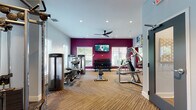 Located at 50 Lily Green Ct NW in Concord, NC, this single-floor fitness center is a well-equipped facility designed to support a comprehensive workout experience. The space is divided into distinct exercise zones, each thoughtfully arranged to maximize functionality and flow.

The cardio area features multiple Life Fitness treadmills, elliptical trainers, and a stair climber, all positioned to allow ample space between machines. A ceiling fan overhead and recessed lighting throughout keep the environment comfortable and well-lit. Motivational wall art and a calming light blue color palette give this section an energizing yet welcoming atmosphere, with natural light filtering in through large windows.

The strength training zone is anchored by a bold magenta accent wall, creating a dynamic visual contrast. A wall-mounted flat-screen TV is centrally positioned, flanked by windows overlooking the outdoors. The area is equipped with a multi-station Life Fitness cable machine, a full dumbbell rack with a wide range of weights, a flat bench, and several selectorized strength machines for targeted muscle training.

Underfoot, striped low-pile carpet tiles run throughout, providing traction and comfort. Storage shelving, a sanitizing station, and exercise balls round out the amenities, ensuring the space is practical and user-friendly. The layout accommodates multiple users simultaneously with ease.