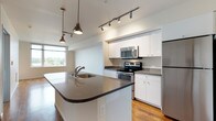 This single-floor residence offers a clean, contemporary design with a thoughtful layout that balances open communal spaces and private retreats. The heart of the home is an open-concept kitchen and living area, unified by warm hardwood-style flooring throughout. The kitchen is well-appointed with white cabinetry, grey countertops, and a full suite of stainless steel appliances, including a range, over-the-range microwave, and refrigerator. A curved kitchen island with a built-in sink and pendant lighting above serves as both a functional workspace and a casual gathering spot. Track lighting illuminates the kitchen area, adding a modern touch. The living area is bright and airy, benefiting from large windows and a glass door that opens to a private balcony, offering pleasant outdoor views and an abundance of natural light. The bedroom features plush grey carpet, neutral white walls, and direct access to a walk-in closet, providing generous storage. The space includes two bathrooms: one with a soaking tub and shower combination, and a second with a glass-enclosed walk-in shower — both finished with wood-look flooring, white vanities, and grey countertops. An additional versatile room with hardwood-style flooring offers flexible use as a home office or den.