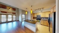 This thoughtfully designed residence showcases an open-concept layout that seamlessly connects the kitchen and living areas. The kitchen is a standout feature, equipped with stainless steel appliances including a refrigerator, range, and microwave, complemented by light maple cabinetry, a tile backsplash, and a generous island topped with dark granite countertops. A pair of elongated amber pendant lights and a track lighting system add both warmth and functionality to the space.

The living area impresses with soaring double-height ceilings, hardwood flooring, and a ceiling fan, while floor-to-ceiling glass doors open to an outdoor balcony, allowing abundant natural light to fill the room. An upper-level loft landing with wood railings overlooks the main living space, adding architectural depth and an airy feel.

The entryway features warm hardwood floors and a staircase with a classic white banister leading to the upper level. Bedrooms are carpeted and bathed in natural light through large windows fitted with horizontal blinds. A bathroom includes tile flooring, granite countertops, and convenient access to a carpeted walk-in closet with built-in shelving and hanging rods. The space offers a clean, modern aesthetic throughout with neutral tones and practical finishes.