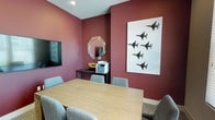 This single-floor communal lounge space offers a well-designed, open-plan layout that seamlessly integrates multiple functional zones. The interior is characterized by a two-tone color scheme, with crisp white walls contrasting against deep slate-blue accent walls, giving the space a contemporary and inviting atmosphere. Light wood-look flooring runs throughout, adding warmth to the modern aesthetic.

The space is divided into distinct yet connected areas. A dining zone features a large wooden table surrounded by bold magenta upholstered chairs, ideal for group gatherings. Adjacent to it, a wall-mounted flat-screen TV is positioned above a paneled accent wall, creating a media-focused lounge area. Further in, high-top bar tables paired with industrial-style adjustable stools provide a casual seating option.

A bar counter area lined with dark bar stools and a mounted TV overhead serves as a social hub, complete with counter space and cabinetry. Decorative wall art featuring a repeating geometric tree motif adds a distinctive visual element throughout. Recessed lighting and a chandelier fixture provide layered illumination, while large windows along the perimeter allow abundant natural light to fill the space.