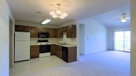 This single-floor residence offers a practical and comfortable layout centered around an open-concept design that seamlessly connects the kitchen and living areas. The kitchen is outfitted with rich, warm-toned wooden cabinetry that provides generous storage space, complemented by light-colored countertops. Essential appliances are included, featuring an electric range, a built-in microwave, a dishwasher, and a sink set conveniently within the cabinetry. A skylight adds a touch of natural brightness to the kitchen area, enhancing the overall sense of openness. An elegant multi-bulb chandelier serves as a focal point, casting a warm and inviting glow throughout the space. The adjacent living area transitions smoothly from the kitchen, featuring plush wall-to-wall carpeting that adds warmth and comfort underfoot. Neutral-toned walls throughout the home provide a clean, versatile backdrop suitable for a variety of interior styles. The open flow between the kitchen and living space creates an inviting atmosphere ideal for both everyday living and entertaining. The overall layout is thoughtfully designed to maximize functionality while maintaining a welcoming and cohesive aesthetic throughout the home.
