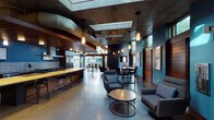 Located at 620 Lenora Street in Seattle's Belltown neighborhood, this single-floor amenity lounge offers a sophisticated blend of industrial and contemporary design. The space is anchored by polished concrete floors, exposed concrete ceilings with visible ductwork, and rich wood paneling that together create a warm yet urban aesthetic. Deep teal accent walls provide a bold backdrop throughout, complemented by carefully curated artwork and pendant lighting that adds a refined ambiance.

The fully equipped kitchen area features stainless steel appliances including double wall ovens and a professional-grade range, dark cabinetry, subway tile backsplash, and a striking butcher-block island with bar seating — ideal for both casual gatherings and culinary events. Adjacent to the kitchen, a generous lounge area is furnished with plush seating arrangements and low coffee tables, encouraging relaxed socializing.

At the far end of the space, floor-to-ceiling glass walls and folding doors open directly onto an expansive outdoor terrace, flooding the interior with natural light and framing panoramic views of Seattle's downtown skyline. The terrace itself offers additional seating for alfresco gatherings. A separate seating nook with a wall-mounted television and direct outdoor access rounds out the versatile layout, making this an exceptionally functional and visually compelling communal space.