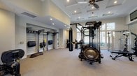 This single-level fitness center is designed to support a comprehensive range of workout disciplines within a spacious, thoughtfully organized layout. The space is divided into distinct functional zones, each tailored to specific training needs.

The main workout area features soaring ceilings accented by a coffered ceiling detail and a ceiling fan, contributing to an open, airy atmosphere. Large floor-to-ceiling windows along one wall flood the interior with natural light and provide views of the surrounding greenery. Carpeted flooring extends throughout, providing comfort underfoot.

At the center of the space, a Hoist Motion cage system serves as the primary multi-functional training station, accompanied by a heavy punching bag and a mini trampoline. A dedicated training zone alcove houses a slatwall storage system stocked with TRX suspension trainers, resistance accessories, and integrated display screens for guided workouts.

The strength training section is equipped with Hoist selectorized machines, a dumbbell rack, medicine balls, plyo boxes, and an adjustable bench. Floor-to-ceiling mirrors visually expand this area while allowing users to monitor form.

Near the entrance, glass double doors provide easy exterior access and additional natural light. A wall-mounted television and ceiling fan complete this welcoming entry zone, making the overall space both functional and well-appointed.