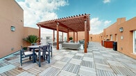 The Sky Lounge at 2422 S Carrier Pkwy in Grand Prairie, Texas is a well-appointed rooftop outdoor amenity space designed for relaxation, socializing, and al fresco dining. The terrace is finished with a distinctive checkerboard pattern of light-toned wood deck tiles, lending a clean and modern aesthetic underfoot throughout the entire level.

At the heart of the rooftop stands a striking large-scale pergola constructed of rich, warm-toned hardwood beams. String lights are woven throughout the pergola's lattice canopy, providing a welcoming ambiance during evening hours. Beneath the pergola, a generous lounge area is furnished with contemporary gray upholstered sofas and accent chairs arranged around low side tables, creating an ideal setting for conversation and casual gatherings.

Adjacent to the covered lounge area, multiple dining sets with round and rectangular tables and coordinating chairs are positioned across the open deck, offering sun-drenched spaces for outdoor meals. Tropical potted plants are strategically placed throughout the terrace, adding a lush, vibrant contrast to the warm terracotta-toned surrounding walls. An outdoor kitchen or grilling station is also visible at the perimeter of the space, further enhancing its functionality as an entertainment destination. Open views of the sky complete the elevated experience of this rooftop lounge.