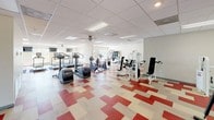 This fitness center offers a spacious, single-floor layout designed to accommodate a comprehensive workout experience. The space is immediately distinguished by its eye-catching geometric floor treatment, featuring a bold red, cream, and gray patterned tile arrangement that energizes the environment and defines the open workout area.

The cardio zone is well-stocked with a row of TRUE-brand treadmills positioned along the perimeter, complemented by elliptical cross-trainers and upright stationary bikes, providing a full range of aerobic exercise options. A dedicated strength-training section houses multiple cable and weight machines, a multi-function cable tower, leg press equipment, and a flat utility bench, catering to users focused on resistance training.

Natural light filters in through windows along the walls, brightening the interior and creating an inviting atmosphere. The drop ceiling with integrated fluorescent panel lighting and ceiling fans ensures consistent illumination and comfortable air circulation throughout. Wall-mounted televisions are also present to keep users entertained during workouts.

Additional amenities include a stability ball and a cubby storage unit for small accessories. The clean, neutral-toned walls and open floor plan contribute to a functional yet welcoming fitness environment suitable for a wide range of exercise routines.