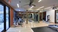 This modern fitness center is thoughtfully designed to support a comprehensive workout experience. The space features light hardwood flooring throughout the main exercise area, complemented by rubber matting zones designated for strength and stretching activities. Warm terracotta-toned accent walls provide a dynamic contrast against neutral columns that are evenly spaced across the floor, adding both structural character and visual definition to the layout. Mirrored walls line multiple sides of the room, creating a sense of expansiveness while allowing users to monitor their form during exercise. Large windows along one wall flood the space with natural light, enhancing the open and energizing atmosphere. Overhead, sleek ceiling fans are suspended from a well-lit ceiling fitted with recessed and linear fluorescent lighting, ensuring bright, even illumination throughout. The cardio section is well-stocked with a row of treadmills, stationary bikes, and elliptical machines, while the strength training area features cable machines, free weights, and resistance equipment. The facility's layout is organized and spacious, allowing for comfortable movement between stations. Overall, the fitness center strikes a balance between functionality and design, offering a clean, professional environment suited for a wide range of fitness routines.