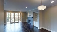 This single-floor space is defined by its refined finishes and thoughtful architectural details. The living area showcases deep, dark-stained hardwood flooring that runs throughout, lending warmth and elegance to the open layout. A central fireplace, clad in light-toned tile, serves as the focal point of the main living space, flanked symmetrically by white built-in shelving and cabinetry units that provide both display and storage opportunities. The surrounding walls are finished in a soft, neutral gray tone that complements the crisp white trim and crown molding, adding a polished, classic character to the interior. Overhead, recessed lighting is supplemented by a stylish drum pendant light fixture, ensuring ample illumination throughout the space. A set of French doors along one wall allows natural light to filter into the room while providing direct access to an outdoor balcony, offering views of the surrounding exterior. The overall ambiance is clean and contemporary, with a balance of functional built-ins, natural materials, and architectural detailing that creates a sophisticated yet inviting living environment.