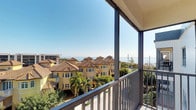 This single-floor space includes a screened balcony that serves as a standout feature, offering an elevated vantage point with expansive, unobstructed views of the surrounding coastal environment. From the balcony, residents can take in a picturesque landscape of Mediterranean-inspired architecture characterized by warm yellow facades and terracotta barrel-tile roofs, interspersed with mature palm trees that lend a tropical character to the setting. Beyond the neighboring rooftops, the shimmering blue waters of the coast stretch to the horizon beneath a clear, open sky, creating a serene and visually striking backdrop. The screened enclosure of the balcony allows for year-round enjoyment of the outdoor space while providing protection from the elements. The elevated positioning enhances both the sense of openness and the quality of the views, making it a natural focal point of the living experience. The overall ambiance is relaxed and resort-like, reflecting the sunny, coastal character of its surroundings and offering an inviting connection between the interior living space and the vibrant outdoor environment.