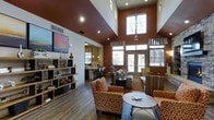 This single-floor space makes an immediate impression with its dramatically high ceilings, which amplify the sense of openness throughout the interior. The design palette centers on deep terracotta and warm rust tones applied to the accent walls, complemented by rich hardwood-style flooring that runs continuously across the space.

The main lounge area is furnished with patterned armchairs and a sofa in coordinating orange and neutral tones, arranged around a round coffee table atop a warm area rug. An open bookshelf along one wall provides both storage and decorative display space, adding a homey, curated quality to the room.

An adjacent bar or counter area is visible toward the rear, featuring dark countertops, bar seating, and pendant lighting suspended from above, suggesting a space suited for socializing or casual dining.

Large windows and glass-paneled entry doors flood the interior with natural light, offering views toward the exterior. A decorative chandelier hangs prominently near the high clerestory window, serving as a central focal point. Recessed lighting throughout further enhances the bright, welcoming ambiance of the space.