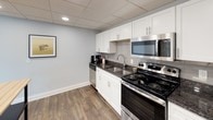 This single-floor amenity space is thoughtfully designed to offer a well-rounded experience across three distinct functional areas. Throughout, a consistent design palette ties the space together, featuring wood-look flooring, drop tile ceilings with large panel lighting fixtures, and light gray walls that create a clean, modern atmosphere.

The lounge area serves as the social centerpiece of the space, furnished with a generously sized sectional sofa accented by colorful throw pillows. Large floor-to-ceiling windows frame a direct view of an outdoor pool and patio area, bringing in ample natural light and creating a seamless visual connection to the exterior amenities. A credenza and round coffee table complete the comfortable, relaxed setting.

The kitchen is fully equipped for practical use, featuring white shaker-style cabinetry, dark granite countertops, and a white subway tile backsplash. Stainless steel appliances — including a range, over-the-range microwave, dishwasher, and full-size refrigerator — provide comprehensive cooking capability, making the space well-suited for gatherings or everyday use.

The fitness center is outfitted with a variety of cardio and strength training equipment, including treadmills, an elliptical, and a multi-station weight machine. A bold teal accent wall and floor-to-ceiling mirror enhance the room's energy and sense of depth, while a water cooler adds a practical convenience for users.