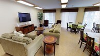 This single-floor communal space is thoughtfully designed to provide comfortable, functional areas for socializing, dining, and everyday living. The layout encompasses a spacious lounge area, a full kitchen, and dedicated dining spaces, all unified by neutral beige walls, plush carpeted flooring, and warm-toned wood accents throughout.

The lounge area is generously sized, furnished with sage green upholstered sofas and armchairs arranged around polished wood tiered coffee tables. A wall-mounted flat-screen television sits atop a classic wood media console, flanked by subwoofer speakers. Wood-framed flush-mount light panels provide consistent, warm overhead illumination across the ceiling. Large windows dressed with vertical blinds and dark valances allow natural light to filter in while offering privacy.

The kitchen features rich medium-toned wood cabinetry, granite-style countertops, and white appliances including a wall oven, microwave, dishwasher, and refrigerator. A pass-through counter connects the kitchen to the adjacent common area, enhancing accessibility and visibility.

The dining area accommodates multiple tables and dark wood chairs, with sliding glass doors offering views of an outdoor patio and lush greenery. A larger communal dining room nearby provides additional seating for group gatherings, lit by matching overhead panel fixtures.