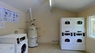 This single-floor laundry facility is designed with practicality and efficiency in mind. The room features a vaulted ceiling that adds a sense of openness, illuminated by overhead fluorescent lighting that ensures a bright, well-lit environment. The walls are finished in a neutral beige tone, contributing to a clean and utilitarian ambiance.

The space is equipped with multiple commercial-grade Speed Queen washing machines and stackable dryer units, providing ample capacity for shared use. The machines are arranged along the walls, maximizing the usable floor space at the center of the room. Instructional posters are prominently displayed on the wall above the washers, offering clear guidance on washing and drying procedures, as well as stain removal tips and service contact information.

A water heater is also housed within the space, integrated discreetly into a corner of the room. The flooring appears to be a durable, speckled finish suited for a high-traffic utility environment. Overall, this laundry facility is thoughtfully organized to deliver a straightforward and accessible experience for its users.