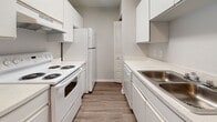 Located at 10700 Buckingham Ct in Port Richey, Florida, this single-story home presents a clean, functional layout with consistent wood-look flooring throughout. The open living and dining area provides a generous sense of space, with a textured ceiling, ceiling fan, and a sliding glass door that invites natural light and provides outdoor access. An additional room, also equipped with a ceiling fan and sliding glass door, offers flexible use as a bedroom or secondary living space.

The galley-style kitchen is efficiently designed with white cabinetry lining both walls, white appliances including a range and refrigerator, a double-basin sink, and light countertops. Adjacent to the kitchen is a built-in desk or utility nook, adding practical workspace to the layout.

Bedrooms feature wood-look flooring, textured ceilings, and windows with blinds that allow for natural light and privacy. Two full bathrooms are present, each finished with white cabinetry, a single-basin vanity with light countertops, a tub-shower combination with tiled surround, and wood-look flooring. The overall palette of neutral grays and whites creates a clean, move-in-ready atmosphere throughout the home.