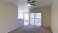 This single-story space features an open-concept layout that connects the living area to a dining space through a pass-through counter, creating a functional and cohesive flow. The living room is carpeted and anchored by a ceiling fan with an integrated light fixture, providing both comfort and overhead illumination. Vertical blinds line the sliding glass door, which opens to an outdoor area visible through the window, offering access to natural light and exterior views. Adjacent to the living area, the dining space benefits from a pendant light fixture suspended from the ceiling, complementing the natural brightness brought in by a large window. The pass-through counter between the dining and kitchen areas enhances the open feel of the space while maintaining a practical separation between zones. Neutral-toned walls and carpeting throughout contribute to a clean, understated aesthetic. The overall ambiance is light and airy, with multiple windows ensuring consistent natural light across the connected living and dining spaces.