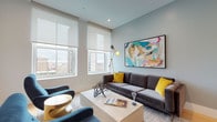 This thoughtfully designed single-floor residence showcases a refined contemporary aesthetic with warm hardwood flooring, soft blue-grey accent walls, and an abundance of natural light streaming through oversized windows that frame sweeping urban skyline views. The floor plan, as revealed in the 3D overview, accommodates two distinct living areas alongside two bedrooms and multiple bathrooms, creating a well-organized and spacious layout.

The primary living area features generous proportions, large roller-shade windows overlooking the cityscape, a charcoal sectional sofa accented with mustard and teal cushions, and a vibrant large-scale abstract artwork. A second, more relaxed lounge area includes a wall-mounted flat-screen TV above a rich wood media console. The kitchen is equipped with dark lower cabinetry, white upper cabinets, a dramatic marble-look backsplash, white quartz countertops, and integrated Smeg appliances.

The bedrooms are serene and well-appointed, with city views, curated artwork, and walk-in closet access. Bathrooms are elegantly finished — one features floor-to-ceiling stone-look tiles with a frameless glass shower, another offers a soaking tub with matching tile surround, and a powder room showcases bold black-and-white hexagonal floor tiles with a floating vanity and round mirror.