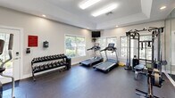 Located at Olde Eastwood Village in Asheville, NC, this fitness center is a well-appointed exercise space designed to support a comprehensive workout routine. The room features durable rubber flooring throughout, providing a safe and functional surface suited for a wide range of activities.

Cardio equipment is a highlight of the space, with two treadmills positioned in front of a bank of large windows that frame views of the surrounding greenery, creating an inviting and naturally lit environment for exercise. A wall-mounted television is positioned above the windows for added entertainment during workouts. An elliptical trainer rounds out the cardio offerings.

Strength training is well represented with a multi-station cable machine, an adjustable weight bench, and a full rack of dumbbells spanning a wide range of weights. A Concept2 rowing ergometer adds a versatile full-body conditioning option to the lineup.

A floor-to-ceiling mirror spans an entire wall, visually expanding the space and allowing users to monitor their form. Recessed lighting and overhead fluorescent fixtures ensure the room is consistently well-lit. French doors provide access to an adjoining area. The overall design is clean and functional, with neutral gray walls and white trim contributing to a bright, focused atmosphere conducive to fitness.