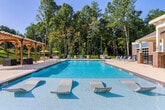 Located along Newnan Crossing Bypass in Newnan, Georgia, this single-story clubhouse serves as a well-appointed community amenity center combining sophisticated interior design with resort-style outdoor features. The exterior presents a classic brick-accented façade with a covered patio area equipped with ceiling fans, offering a shaded retreat directly adjacent to the sparkling pool. The expansive swimming pool is surrounded by a wide, open sundeck furnished with lounge chairs, ideal for relaxation under Georgia's open skies. A separate accessory structure near the pool provides additional support space for outdoor activities.

Inside, the main lounge is distinguished by soaring ceilings, warm hardwood-style flooring, and dramatic dark accent walls adorned with decorative molding panels. A statement chandelier anchors the seating area, which features contemporary furnishings arranged around a central coffee table. Large windows flood the space with abundant natural light. An adjacent dining and social area continues the dark, modern aesthetic with bistro-height tables, bar stools, decorative wall art, and a mounted flat-screen television. A kitchenette area rounds out the interior, offering granite countertops, dark cabinetry, stainless steel appliances, and a mounted display screen, completing this thoughtfully designed community gathering space.