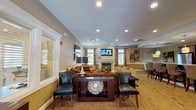 Located at 110 Westwood Dr in Westbury, NY, this single-floor community clubhouse offers a well-appointed gathering space designed for comfort and active living. The lounge area serves as the heart of the facility, featuring a classic white-mantled fireplace framed by large windows dressed with elegant drape curtains, allowing generous natural light to fill the room. Warm-toned walls, recessed lighting, and wood-style flooring create an inviting ambiance, complemented by plush seating arrangements and wall-mounted flat-screen televisions. A dining nook adjacent to a kitchenette includes a dark wood table, upholstered seating, a stainless steel refrigerator, and rich wood cabinetry, providing a functional space for casual meals or gatherings. The expansive fitness center is a standout feature, offering a comprehensive selection of Life Fitness cardio equipment including ellipticals, treadmills, and stationary bikes, all positioned alongside large windows that overlook the surrounding greenery. Strength training is well accommodated with cable machines, a multi-function weight station, a dumbbell rack, and a workout bench. Built-in water fountains and wall-mounted televisions further enhance the fitness experience, making this clubhouse a complete and practical community amenity.