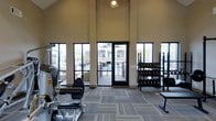 This fitness center offers an open, airy workout environment defined by its impressively high ceilings and expansive floor-to-ceiling windows that flood the space with natural light. The large black-framed windows and central glass door provide views of the surrounding exterior and offer direct access to an outdoor area, creating a seamless indoor-outdoor connection. The neutral-toned walls complement the modern patterned carpet flooring, which adds texture and a contemporary aesthetic to the space. The facility is well-equipped with a range of strength training and cardio equipment, including a multi-function cable machine and a leg press or extension machine visible on one side, while a full dumbbell rack with an accompanying flat bench anchors the free weights area on the opposite side. The layout is thoughtfully organized to allow comfortable movement between equipment. A ceiling-mounted light fixture provides additional ambient lighting, enhancing the bright atmosphere created by the generous window placement. Overall, the fitness center presents a clean, modern, and functional environment well-suited for a comprehensive workout routine.