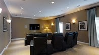 This single-floor home theater space is thoughtfully designed to deliver a true cinematic experience. The room is anchored by multiple rows of plush, oversized black leather recliner chairs arranged in a tiered layout, ensuring unobstructed sightlines to the large wall-mounted flat-screen television at the front of the room. The warm, tan-toned walls are complemented by elegant crown molding and a combination of recessed ceiling lights and wall sconces, which create a soft, ambient glow ideal for movie viewing. A decorative table lamp adds an additional layer of warmth to the room's atmosphere. Classic Hollywood-inspired framed artwork adorns the walls, reinforcing the theater aesthetic. A media console positioned beneath the television neatly houses audio and visual equipment. A window dressed with floor-length curtains allows for natural light when desired, while maintaining the room's intimate ambiance when drawn. The neutral carpeted flooring ties the space together, absorbing sound and enhancing the acoustic quality of the room. Overall, this home theater offers a well-appointed, comfortable, and stylish retreat designed for entertaining and immersive film experiences.