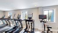 Located on Rolling Green Dr in Milford, MA, this fitness center offers a well-appointed, single-floor space designed to support a comprehensive workout routine. The room is finished with a stylish patterned carpet in neutral tones that adds texture underfoot while maintaining a clean, contemporary aesthetic throughout.

The cardio area is prominently featured and houses a lineup of modern machines, including treadmills, ellipticals, and a stationary cycling bike, each equipped with individual digital display screens for an interactive exercise experience. A recumbent bike provides an additional low-impact cardio option. The strength training section features a multi-function cable machine along with a dedicated bench, offering versatility for resistance-based workouts.

Natural light filters in through multiple windows, creating a bright and inviting atmosphere. Recessed ceiling lighting supplements the natural light, ensuring the space remains well-lit at all hours. A striking dark blue glazed tile accent wall adds a bold design element and visual depth to the room, contrasting elegantly with the soft gray walls.

Practical amenities include a dual-level water fountain station, a fire extinguisher, and posted safety information. Glass-paneled doors provide access to adjacent areas of the building, contributing to an open, connected feel. The overall ambiance is clean, modern, and purposefully designed to support an active lifestyle.