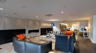 This single-floor space showcases a sophisticated contemporary design anchored by a striking floor-to-ceiling stone fireplace wall with a wide linear gas fireplace as its centerpiece. The open-concept layout seamlessly connects the main lounge area to an adjacent room visible in the background, creating a sense of generous, flowing space.

The main living area is furnished with a combination of deep charcoal leather seating and plush blue velvet armchairs, accented with vibrant orange throw pillows that add bold contrast to the neutral palette. A rectangular glass-top coffee table sits atop a subtly patterned area rug, completing the central seating arrangement.

Recessed lighting is distributed evenly across the smooth white ceiling, supplemented by a modern arc floor lamp with a blue shade that adds both ambiance and functional task lighting. The walls feature a clean, minimalist finish that complements the stone fireplace surround.

The adjoining room, partially visible through an open passage, appears to offer additional seating and natural light, suggesting a versatile, multi-functional layout. The overall ambiance is polished, modern, and inviting.