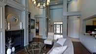 This single-floor space makes a striking impression through its dramatic double-height ceilings, which lend an expansive, airy quality to the main living area. Towering decorative columns finished in soft gray anchor the room, framing the space with architectural distinction and adding a sense of formality without sacrificing warmth. A classic wrought-iron chandelier with cylindrical shades hangs prominently overhead, serving as a focal point that draws the eye upward and reinforces the room's grand scale.

The interior palette is composed of cool, muted gray tones across the walls, millwork, and built-in cabinetry, creating a cohesive and refined aesthetic throughout. Dark hardwood flooring grounds the space and provides rich contrast against the lighter wall treatments and furnishings. A tiled fireplace on one wall adds a traditional element and sense of comfort to the living area. Seating includes a plush gray sofa and a crisp white accent chair, complemented by a glass-and-metal coffee table that adds a contemporary touch.

Toward the rear, a set of glass-paneled double doors floods the space with natural light and offers views of the surrounding exterior, connecting the interior to the outdoors. Additional rooms are accessible through framed doorways, suggesting a well-connected and thoughtfully designed floor plan.