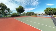This outdoor tennis court offers a well-maintained hard-surface playing area finished in a classic combination of green and terracotta red, with crisp white boundary and service lines clearly marked throughout. The court is fully enclosed by chain-link fencing, providing both safety and visibility while maintaining an open, airy feel. The net is properly installed at regulation height, making the space ready for competitive or recreational play.

The setting is notably scenic, with the court positioned adjacent to a calm body of water that reflects the surrounding landscape. Mature, leafy trees frame the perimeter, adding natural shade and a sense of seclusion. Neatly trimmed hedgerows line portions of the fencing, contributing to the well-kept aesthetic of the space.

Open to a wide expanse of sky, the court benefits from abundant natural light throughout the day. The combination of the waterfront view, verdant greenery, and spacious playing surface creates an inviting and visually appealing recreational environment. The overall ambiance is tranquil yet active, making this court a standout amenity suited for players of all skill levels.