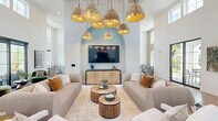 Located at 1450 Front Place in North Port, Florida, this thoughtfully designed single-story clubhouse offers a generous and versatile gathering space for its community. The entrance welcomes visitors with light wood flooring, black-framed glass doors, and an open, airy feel that sets the tone throughout.

The main living area is the centerpiece of the clubhouse, boasting dramatic soaring ceilings lined with transom windows that flood the space with abundant natural light. A cluster of woven rattan pendant lights hangs overhead, adding a warm coastal aesthetic. Comfortable neutral-toned sofas and lounge chairs are arranged around a built-in arched accent wall housing a large flat-screen television, creating an inviting social hub.

Adjacent to the lounge is a stylish bar and dining area featuring a striking dark marble-topped island with upholstered seating, sage green cabinetry, a mosaic-tiled arched backsplash, and globe pendant lighting — all set on polished terrazzo flooring.

The game room area provides additional recreational space, complete with a full-size billiards table and panoramic windows offering views of the surrounding Florida landscape, including tropical palms. Private office or meeting rooms with black-framed glass walls offer functional workspace within the facility. Throughout, the design blends coastal elegance with modern sophistication.