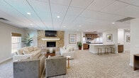 Located at 3300 Montavesta Rd in Lexington, KY, this well-appointed clubhouse offers a thoughtfully designed layout across a generous single floor. The living room serves as the heart of the space, anchored by a dramatic floor-to-ceiling natural stone fireplace with a white mantel and wall-mounted TV, flanked by comfortable seating arrangements atop patterned carpet. Large windows dressed with woven wood shades flood the area with natural light and offer views of the outdoor pool and surrounding greenery. The open-plan kitchen is outfitted with rich walnut cabinetry, stainless steel appliances, white quartz countertops, and light hardwood flooring. A wide peninsula with bar seating provides an inviting gathering spot. Adjacent to the main living area, a separate room features a full-size billiards table, adding a recreational dimension to the space. Glass-paned French doors connect the various zones, maintaining an open and airy feel throughout. A secondary lounge area with natural light and outdoor views offers additional seating for smaller gatherings. Two bathrooms and recessed lighting throughout complete this versatile, welcoming space, all finished with a drop ceiling and neutral tones that lend a clean, modern ambiance.