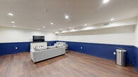 Located at 1960 PA-611 in Swiftwater, Pennsylvania, this single-floor space offers a cohesive and thoughtfully designed interior centered around a spacious living room. The room is immediately distinguished by its striking two-tone wall treatment, featuring crisp white upper walls paired with bold navy blue lower wainscoting that runs throughout, lending the space a sharp, modern aesthetic.

Rich, dark hardwood-style flooring flows across the entire floor, providing a warm contrast to the cool color palette above. Recessed lighting is evenly distributed across the white ceiling, delivering bright, even illumination throughout. The living area is furnished with comfortable gray sofas, accent pillows, and a low-profile coffee table, all arranged around a large flat-screen television mounted atop an ornate white credenza.

A fully equipped kitchen area is also present, outfitted with navy blue cabinetry that mirrors the wall accents, stainless steel appliances including a refrigerator and range, a stainless steel wall-mounted range hood, and a sink with counter space. Glass double doors connect this kitchen area to an adjacent corridor, adding both accessibility and openness to the layout.

The overall ambiance is clean, polished, and functional, making this space well-suited for gathering and everyday use.