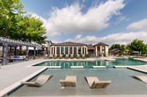 Located at 605 Winterfield Dr in Hutto, Texas, this single-level outdoor amenity space serves as a resort-style retreat within a residential community. At its centerpiece is a large, freeform swimming pool with a striking organic shape, complemented by a separate attached spa for relaxation. The expansive pool deck is finished in a light-toned aggregate surface and is accessible via stone-accented stairways framed by native landscaping, including palm plants, manicured hedges, and decorative river rock beds. Ample lounge seating and sun chairs are arranged around the pool perimeter, offering plenty of space for sunbathing and outdoor gatherings. A second rectangular pool area with in-water lounge ledges provides an additional swimming zone. Shaded areas are provided by a modern pergola structure and a covered outdoor seating area equipped with a fireplace, ideal for social gatherings. A dedicated pool house with large screened doors offers functional support facilities. Mature trees provide natural shade throughout the grounds, while well-maintained landscaping enhances the overall ambiance. The space is enclosed by decorative metal fencing, ensuring privacy and security. Open skies and views of the surrounding neighborhood complete this well-appointed outdoor community amenity.