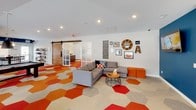 This single-floor community room is a well-appointed shared space designed for both socializing and relaxation. The layout is open and expansive, allowing multiple activity zones to coexist comfortably within one cohesive environment. A striking hexagonal carpet pattern in shades of red, orange, and neutral tones anchors the space visually and adds a bold, modern character underfoot.

The lounge area features multiple sectional sofas arranged to encourage conversation, complemented by a wall-mounted fireplace set against a wood-paneled accent wall and a deep teal blue accent wall. A wall-mounted television, decorative wall art, and a barn-style sliding door add personality and functional versatility to the room. Natural light filters in through multiple windows fitted with horizontal blinds, providing outdoor views and brightening the interior.

A games area includes a pool table and billiard cue rack, offering recreational options for residents. The kitchen zone is fully equipped with dark cabinetry, stainless steel appliances, and a long bar-height counter with seating for several guests. A warm wood-planked ceiling above the kitchen area adds texture and visual warmth, while pendant lighting illuminates the bar counter below.