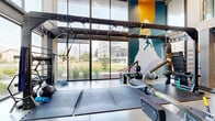 This expansive fitness center offers a comprehensive workout environment housed within a single open floor plan of generous proportions. The space features an industrial-modern design aesthetic, with exposed ductwork, structural beams, and sleek linear LED lighting running across the ceiling, creating a dynamic and energetic atmosphere throughout.

The facility is divided into distinct training zones. A dedicated strength area houses a wide array of selectorized cable machines and plate-loaded equipment from True Fitness, complemented by a full rack of dumbbells and multiple weight benches. Flooring transitions between polished concrete and rubber matting to suit each zone's function. The cardio section features a row of treadmills and stair climbers positioned directly against floor-to-ceiling glass windows, providing ample natural light and open views of the surrounding outdoor environment.

A functional training zone includes a heavy bag, overhead rig, plyo boxes, battle ropes, and medicine balls, suitable for high-intensity and cross-training workouts. Full-length mirrors line key sections of the walls, enhancing the sense of space. Vibrant, large-scale fitness-themed murals add bold color and artistic character to the interior. The facility is climate-controlled with a visible HVAC system and benefits from abundant natural light streaming through expansive window walls.