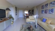 This single-floor residence offers a cohesive open layout connecting the living and dining areas in a seamless flow. The living area is anchored by a pair of cream-colored sofas arranged around a glass coffee table, accented by decorative candles and a tall floral arrangement that adds an elegant touch. A patterned area rug defines the seating space atop the light-toned tile flooring, which extends throughout the entire unit, contributing to a clean and airy aesthetic. Warm beige walls are complemented by framed artwork and a coastal-themed canvas, lending the space a relaxed yet refined character. A tall potted plant near the entryway adds a natural element to the room. Beyond the living area, the dining space accommodates a wood table with matching chairs, illuminated by a pendant light fixture. A tall, dramatic indoor plant serves as a natural divider between the two zones. Natural light filters in through windows visible at the far end of the dining area, brightening the space. The layout is practical and well-organized, with multiple doorways leading to additional rooms, maintaining an uncluttered and inviting atmosphere throughout.
