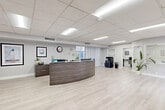 Located at 2903-2915 NW 60th Terr in Sunrise, Florida, this well-appointed single-floor commercial space offers a clean, modern aesthetic designed for professional use. The layout flows seamlessly from a bright, open reception and waiting area near the entrance — furnished with sleek black leather seating, a marble-top accent table, and wall-mounted displays — to a curved, dark wood reception desk that serves as the functional centerpiece of the space. Light wood-look flooring runs throughout, complementing the crisp white walls and drop-panel ceiling with recessed and panel lighting that keeps the entire space evenly illuminated. Large windows and sliding glass doors at both ends of the suite allow generous natural light to pour in and provide views of the surrounding outdoor deck and landscaping. The kitchen area features a white peninsula counter with bar-height stools, ideal for breaks or informal meetings. Two private offices offer dedicated workspaces, while a closet provides additional storage. The overall ambiance is polished and professional, with a neutral color palette and thoughtful layout that balances an open, collaborative feel with practical privacy — making this a versatile and functional commercial environment.