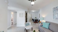 This single-floor residence offers an open-concept layout that connects the living and dining areas in a cohesive, flowing arrangement. The space is finished with light-toned laminate or luxury vinyl flooring throughout, lending a clean and airy feel to the interior. Walls are painted in a soft, neutral white, maximizing the sense of openness and allowing decorative accents in teal and warm tones to stand out effectively.

The living area is anchored by a ceiling fan, providing both functional ventilation and a subtle design element overhead. A cozy accent chair with nailhead trim detailing sits at the edge of the seating area, complementing the grey sofa nearby. The dining zone features a round glass-top table surrounded by dark upholstered chairs, positioned on a area rug that defines the space within the open floor plan.

A hallway visible from the main living area leads toward additional rooms, with a closet and what appears to be a kitchen situated beyond a partial wall. Decorative wall art adds a personal touch to the corridor. Overall, the space presents a tidy, well-coordinated interior with a bright, welcoming ambiance.