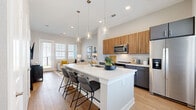 This thoughtfully designed 2-bedroom, 1-bathroom residence makes efficient use of its single-floor layout, offering a bright and modern living experience. The open-concept kitchen and living area serves as the heart of the home, anchored by a large central island with a white quartz countertop, undermount sink, and seating for multiple guests. Pendant lights with glass globe shades hang overhead, while two-tone cabinetry — warm wood uppers paired with charcoal lowers — and a full suite of stainless steel appliances complete the contemporary kitchen design. A crisp subway tile backsplash adds texture to the space. Light hardwood flooring flows seamlessly throughout the main living area, enhancing the sense of openness. The living room receives generous natural light through large windows and a glass-paneled door that opens to an outdoor balcony. Both bedrooms are well-proportioned, with the primary bedroom featuring multi-window exposure, a dedicated walk-in closet with built-in shelving and hanging rods. The full bathroom is finished with large-format tile in the tub/shower combo, a glass sliding door, and a gray vanity with white countertop and brushed nickel fixtures. Neutral gray walls and warm wood tones tie the aesthetic of the entire residence together cohesively.