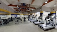 This single-level fitness center is distinctly branded in West Virginia University's signature blue and gold, with "Let's Go Mountaineers" and the WVU flying WV logo prominently displayed throughout the space. The facility's most striking architectural feature is its vaulted ceiling supported by exposed warm-toned wood trusses, which lend a lodge-like character to the interior while creating a generous sense of vertical openness. Visible ductwork, ceiling fans, and pendant lighting are integrated throughout the ceiling structure, ensuring both comfort and adequate illumination across the entire floor.

The fitness floor is divided into distinct functional zones. A dedicated cardio area lines the perimeter walls, where rows of treadmills, elliptical trainers, and stationary bikes are positioned directly in front of expansive windows. These large windows offer views of an outdoor patio and natural surroundings, providing ample natural light and an inviting atmosphere during workouts. A separate strength training zone houses cable machines, adjustable benches, free weight racks with dumbbells, and weight benches, all arranged with sufficient spacing for safe use. Full-length mirrors along one wall enhance the sense of space and assist users during strength exercises. Framed artwork adds a finishing touch to the walls, complementing the overall well-appointed, community-oriented character of the facility.