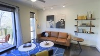 This single-floor space showcases a thoughtfully designed living area that balances modern aesthetics with an eclectic personality. The main living space is anchored by a rich tan leather sofa, positioned atop a bold blue geometric area rug that adds a striking visual contrast to the dark hardwood flooring. A ceiling fan with integrated lighting and a track lighting system provide layered illumination throughout the room, ensuring the space is well-lit and functional.

The walls are adorned with a pair of large-scale black-and-white art prints featuring stylized cat portraits, lending the space an artistic, urban character. A modular open shelving unit along one wall offers both display and storage opportunities. Complementing the seating area are two round white coffee tables and a coordinating side table, creating a cohesive and practical arrangement.

Natural light filters into the space through a floor-to-ceiling window or glass door on one side, offering a connection to the outdoors. A sleek floor lamp adds warm accent lighting, while small decorative objects and books throughout reinforce the curated, contemporary ambiance of this well-appointed living space.