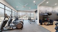 This contemporary fitness center is thoughtfully designed across a single floor, offering two distinct workout zones that together provide a comprehensive exercise experience. The main cardio and strength area is bright and airy, anchored by expansive floor-to-ceiling windows that flood the space with natural light and offer open views of the surrounding outdoor environment. A teal-painted tray ceiling with a large ceiling fan adds a stylish architectural touch, while recessed lighting ensures excellent visibility throughout. This zone is outfitted with treadmills, elliptical trainers, a rowing machine, and adjustable weight benches, complemented by a full rack of dumbbells along the windowed wall. A wall-mounted flat-screen TV is positioned for easy viewing during workouts. The second zone, finished with light hardwood-style flooring, serves as a functional training and studio area. It features floor-to-ceiling mirrors, a cable and weight machine, a spin bike with an interactive display, and an organized selection of resistance training accessories. The transition between zones is defined by a structural column with built-in storage cubbies and an additional wall-mounted display screen. Together, both areas deliver a well-rounded, modern fitness facility suitable for a wide range of training styles.