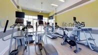 This fitness center offers a bright, energizing environment designed to support a well-rounded workout routine. The space is finished with light hardwood-style flooring and yellow-accented walls accented by a neutral horizontal stripe, creating a vibrant yet focused atmosphere. A prominent "fit·ness" wall graphic reinforces the space's dedicated purpose.

The cardio area is well-stocked with a variety of equipment, including treadmills, elliptical trainers, stair climbers, and stationary cycling bikes. Each machine is positioned to allow comfortable spacing, and several units are equipped with individual monitors for entertainment during workouts. Additional wall-mounted flat-screen TVs are positioned throughout the room for easy viewing from multiple stations.

Natural light flows generously into the space through large floor-to-ceiling windows and glass doors along one wall, offering views of the outdoors and enhancing the open, airy feel of the room. A ceiling fan overhead contributes to air circulation and comfort during active use. Recessed and overhead lighting supplements the natural light, ensuring the space remains well-lit throughout the day. Overall, the fitness center is thoughtfully designed to provide a motivating and functional environment for residents.