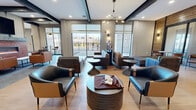 This expansive amenity space combines a sophisticated lounge with a fully equipped game room, all unified by a modern industrial aesthetic. The single-floor layout flows seamlessly between two distinct zones, each thoughtfully designed for socializing and recreation. Exposed dark steel beams contrast beautifully against neutral gray walls and warm light wood flooring, establishing a refined yet inviting atmosphere throughout.

The lounge area features multiple seating arrangements, including leather and upholstered chairs grouped around sculptural pedestal tables, bathed in natural light from floor-to-ceiling windows that offer outdoor views. A striking multi-arm pendant chandelier and recessed lighting provide layered illumination. A tiled linear fireplace anchors one wall, accompanied by a wall-mounted TV and warm leather seating, creating an intimate gathering zone. Decorative chain-link mesh panels and reclaimed wood accent walls add unique industrial character.

The game room is generously proportioned to accommodate a full-size pool table, a shuffleboard table, and a ping-pong table, all set on gleaming hardwood floors. Pendant lighting and curated wall art complete the space. Large windows and glass doors connect both areas visually to an outdoor courtyard, ensuring an abundance of natural light and a sense of openness throughout.